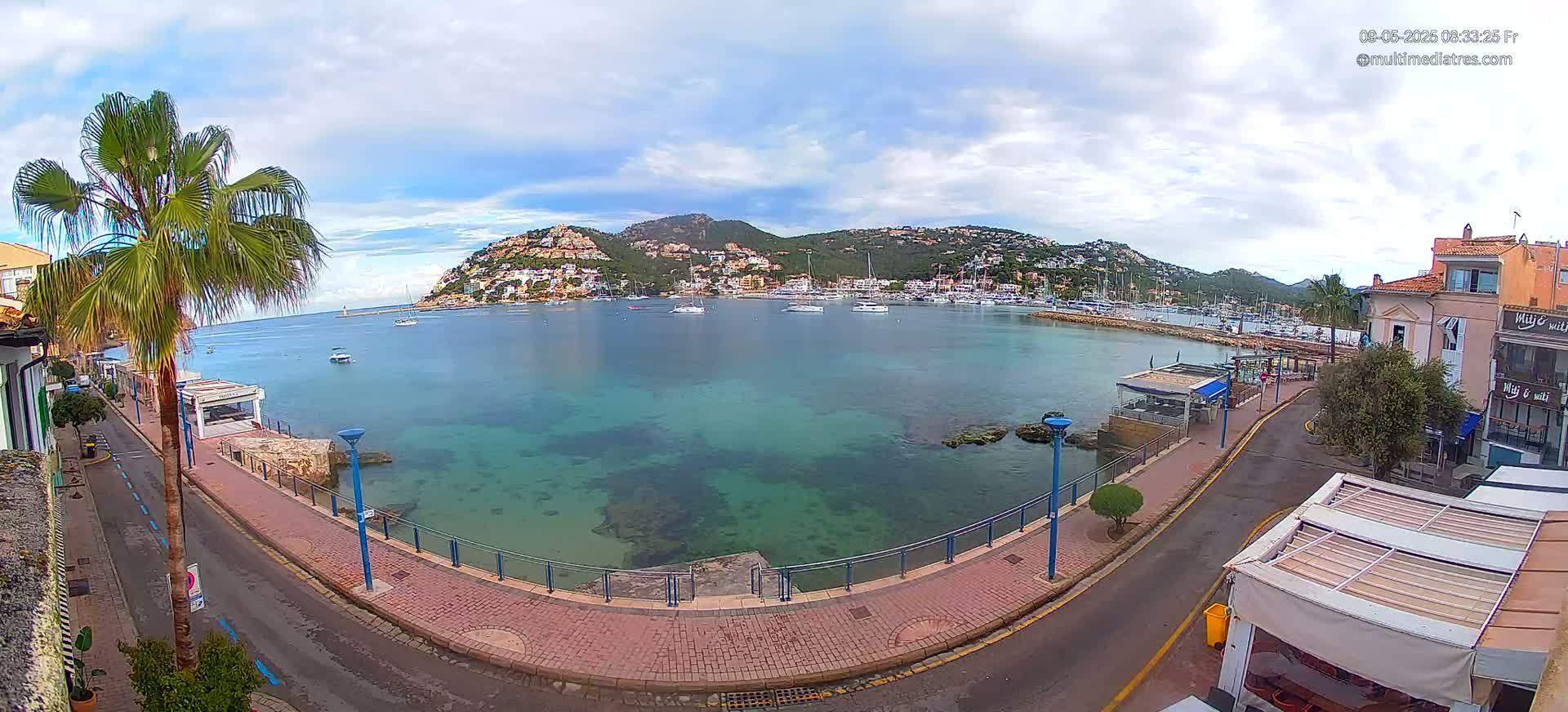 A partly cloudy day overlooking a calm, clear, turquoise bay with moored boats, a town, and hills in the background.