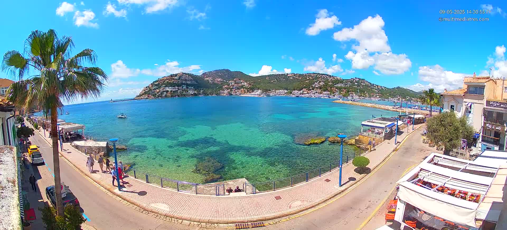 A sunny day shows a clear, turquoise bay with boats and a hillside town, a palm tree, a paved waterfront walkway, and several people walking along the street.
