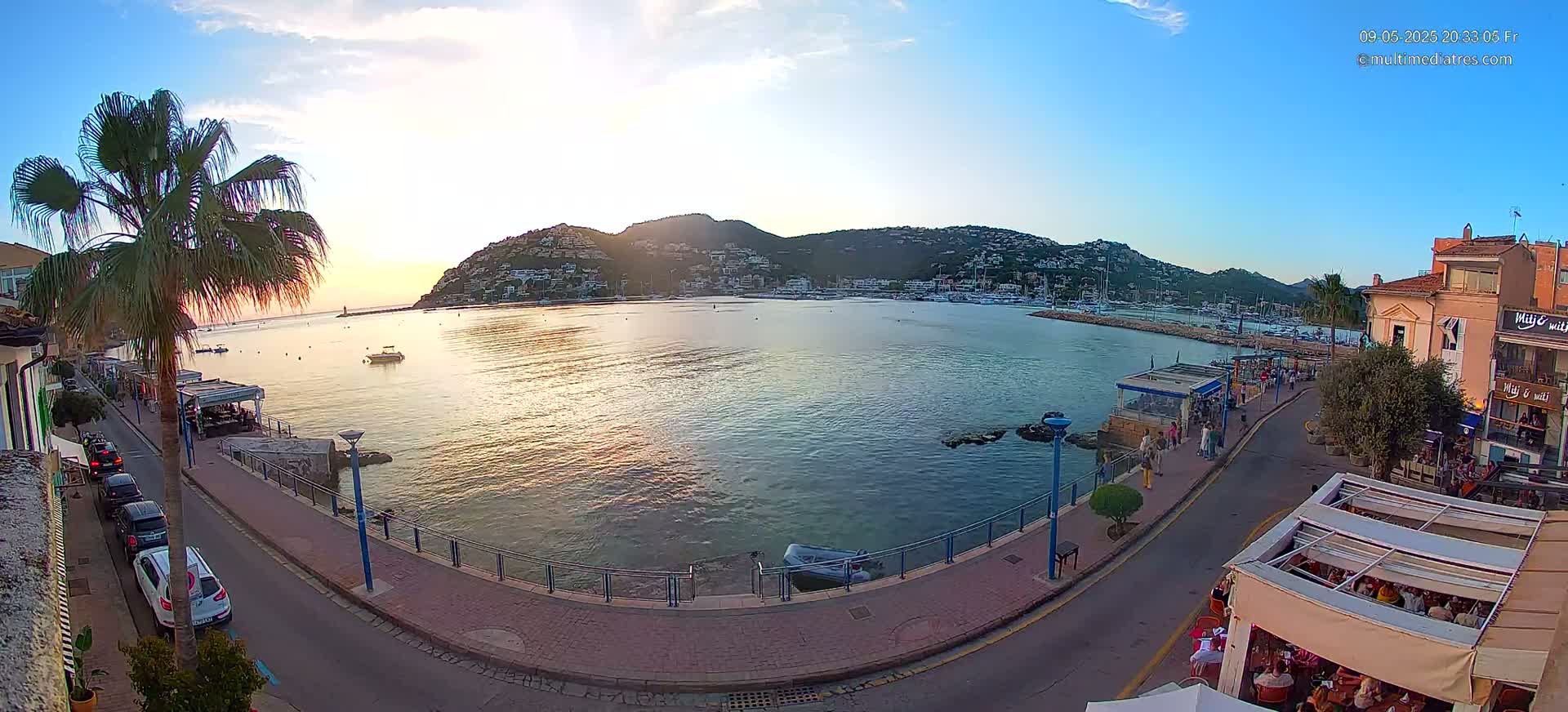 A seaside town at sunset, with calm water reflecting the golden light,  cars parked along a paved waterfront path lined with restaurants, and a hill covered in buildings in the background.