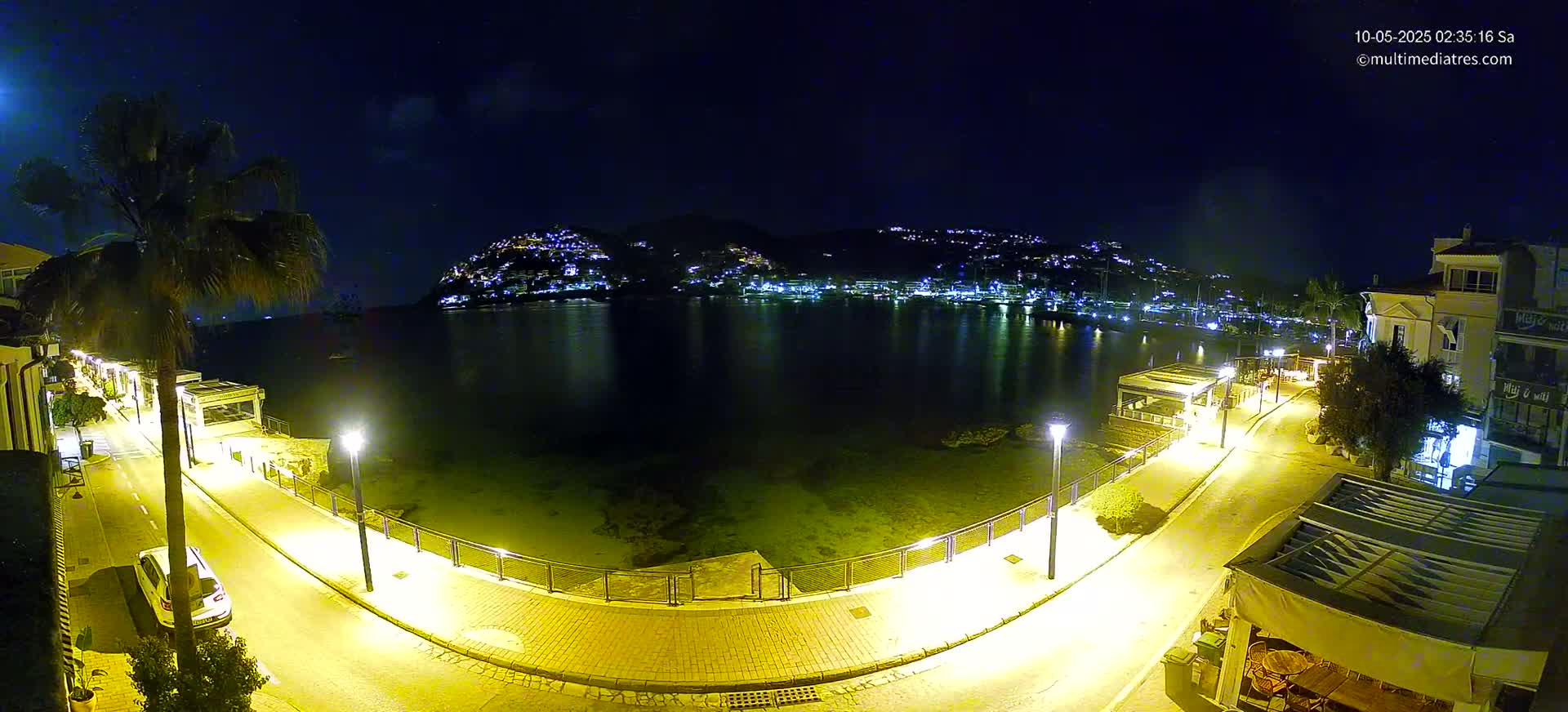 A nighttime, illuminated waterfront scene shows a calm bay reflecting the lights of buildings on a hillside across the water, with a paved walkway and streetlights lining the shore.