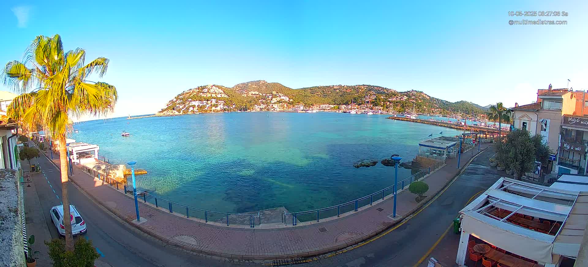 A clear, sunny day overlooks a calm, teal-colored bay with a town nestled on a hillside in the background, seen from a waterfront street with a palm tree and parked car.