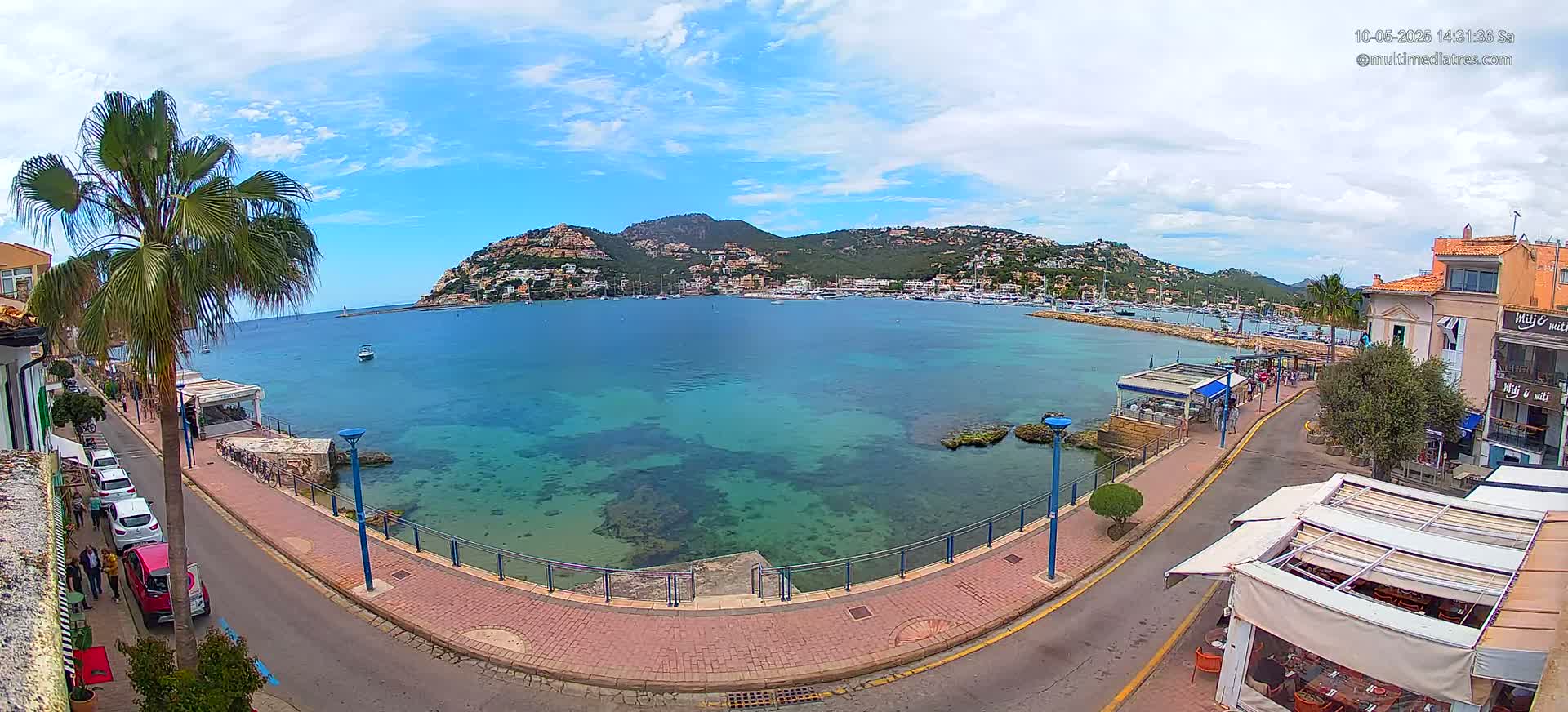 A partly sunny day shows a clear, turquoise bay with a town and hills in the background;  a paved walkway with parked cars lines the water's edge.