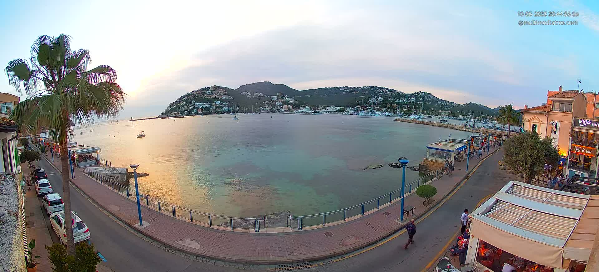 A calm bay with boats at sunset is viewed from a waterfront promenade with parked cars and people walking by restaurants.
