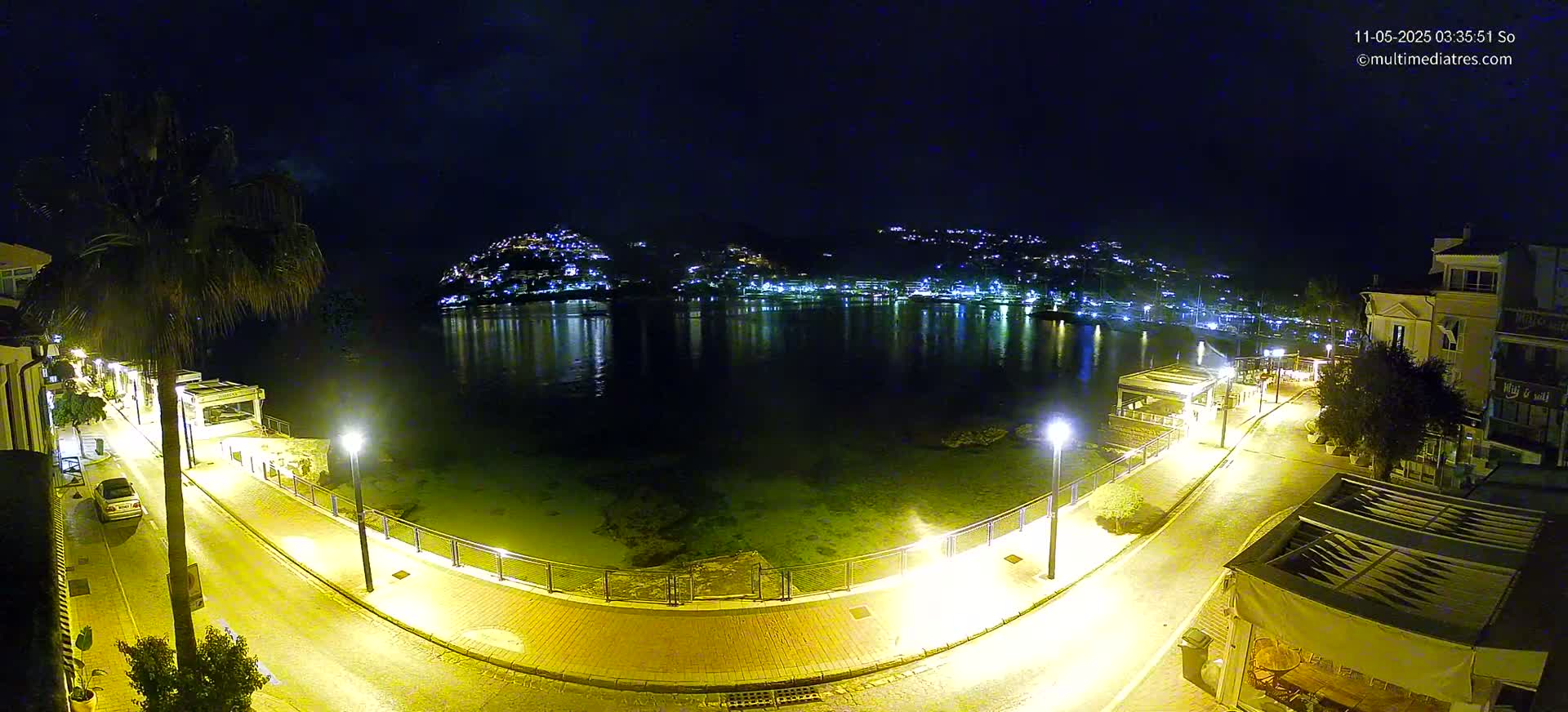 A nighttime, brightly lit waterfront scene shows a calm bay reflecting the lights of a hillside town, with a paved walkway curving alongside the water's edge.