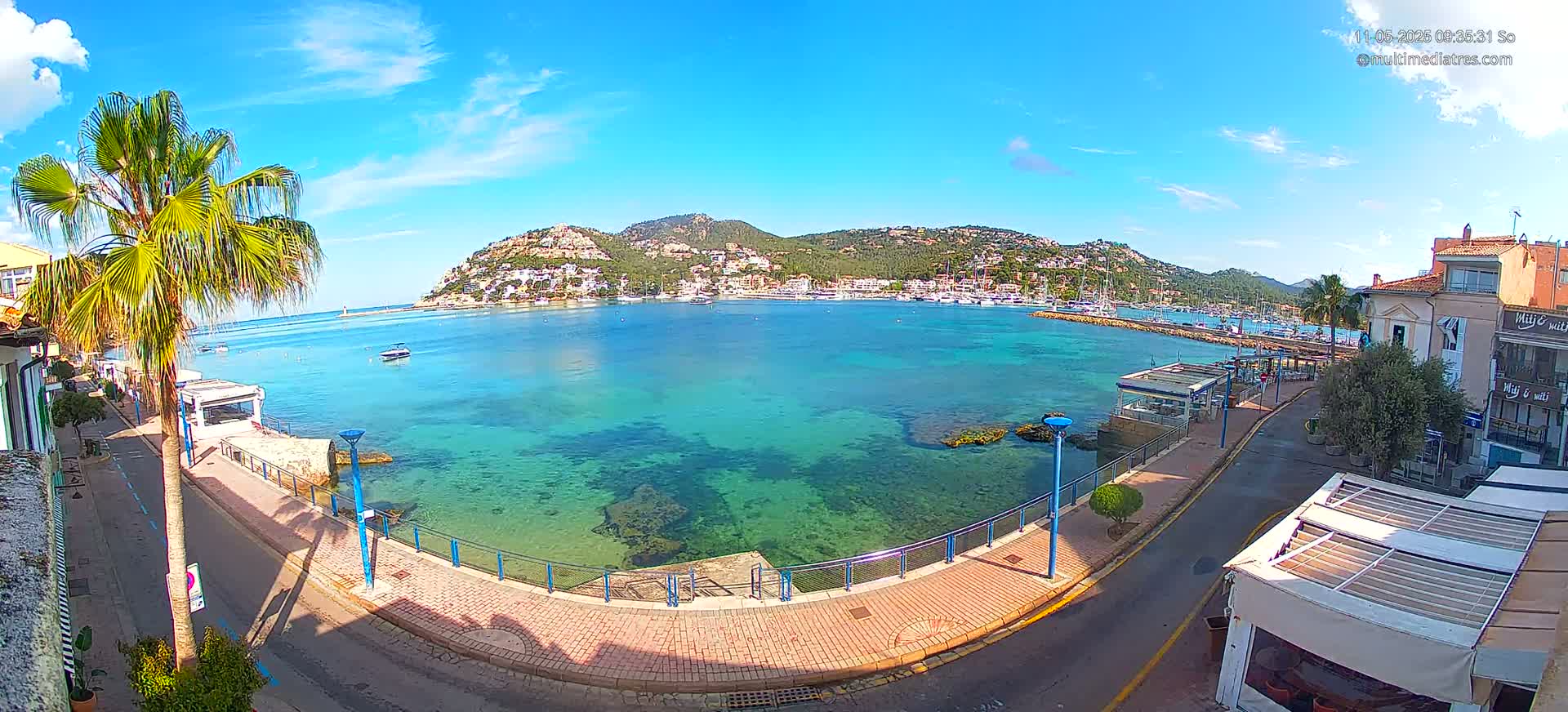 A clear, sunny day overlooking a calm, turquoise bay with a town and hills in the background, viewed from a seaside promenade.