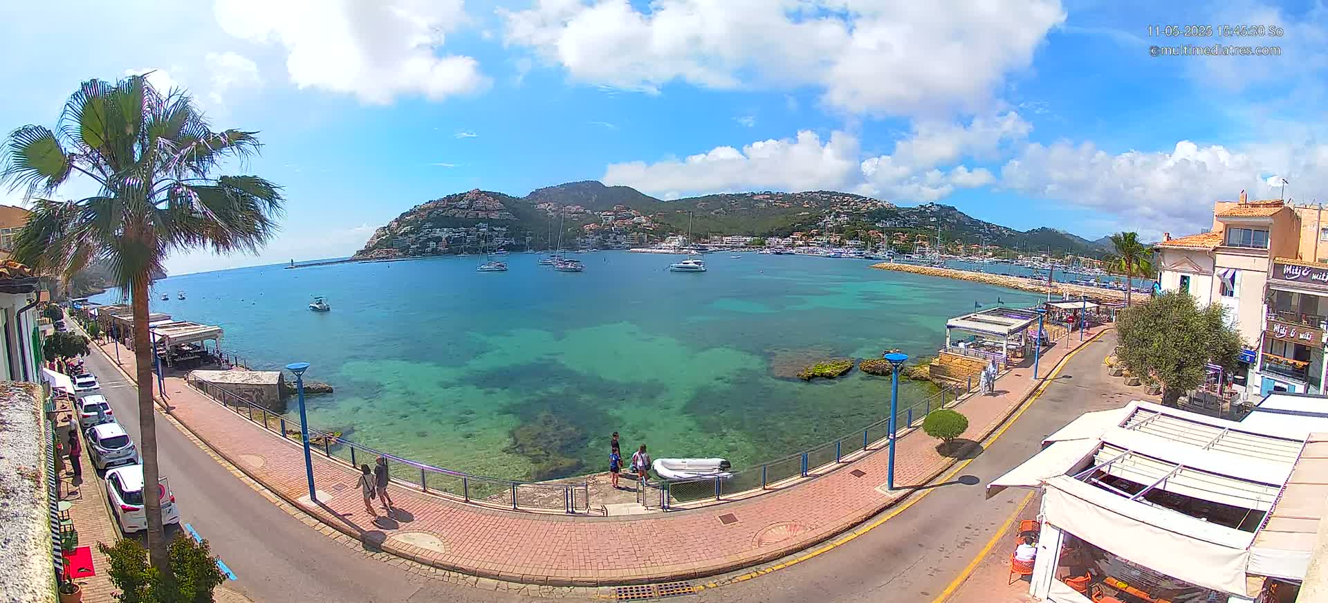 A sunny day reveals a picturesque coastal town with a calm, clear turquoise bay filled with boats, a paved waterfront walkway, and buildings lining the shore.
