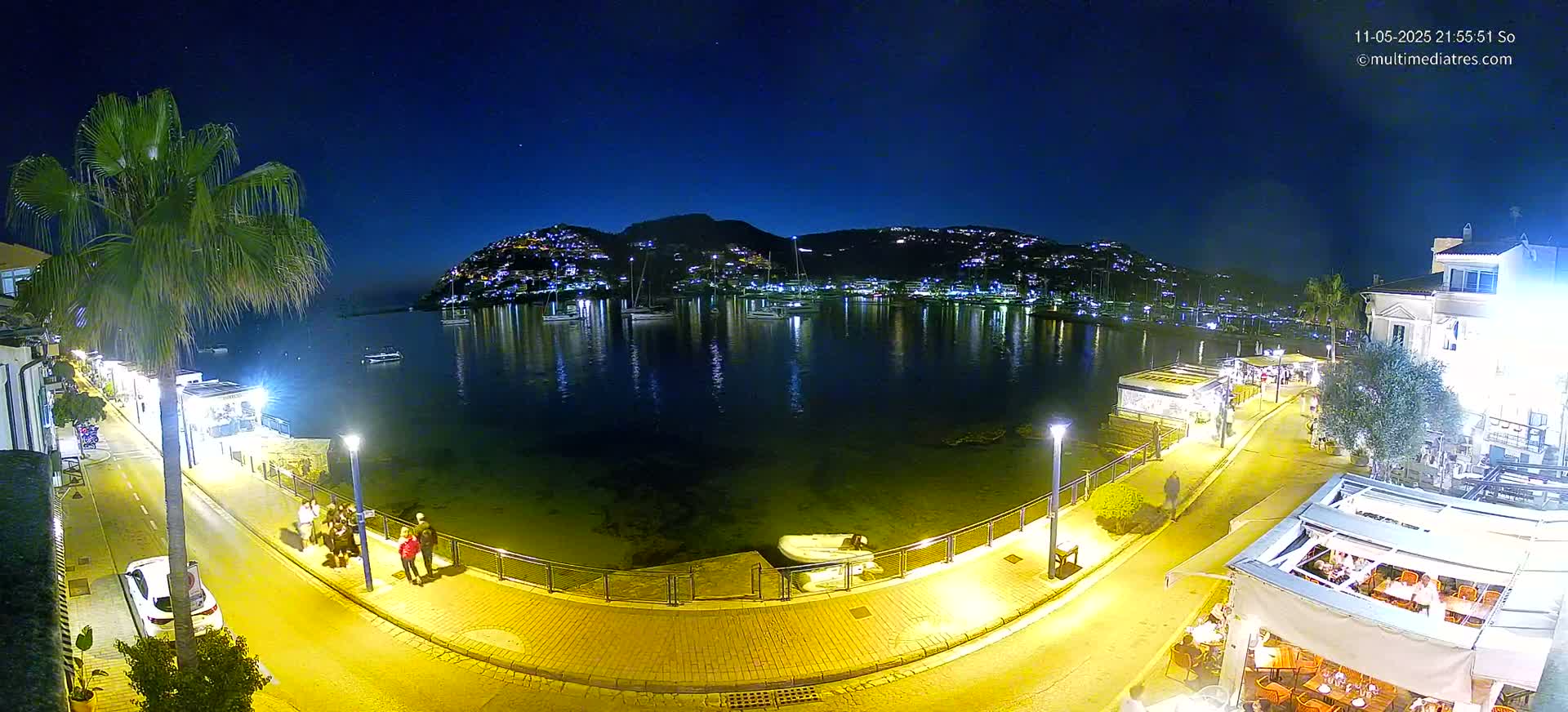 A nighttime harbor scene with calm water reflecting the lights of buildings and boats on a hill, illuminated by streetlights along a waterfront walkway where people are strolling.
