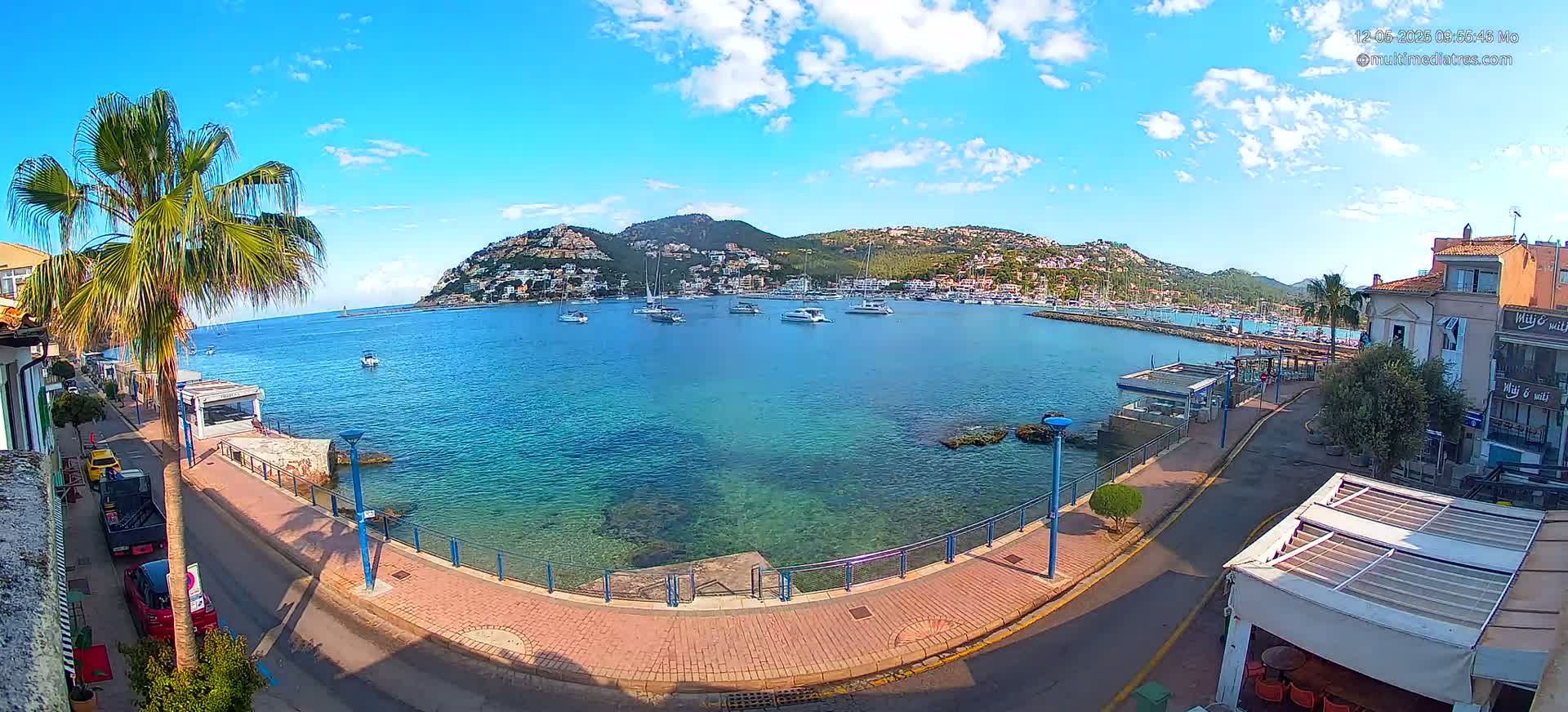 A seaside town with clear blue water, numerous boats in a harbor, and buildings lining a paved walkway under a mostly sunny sky.