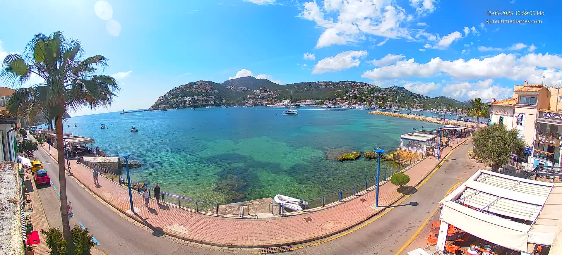 A sunny day overlooks a clear, calm bay with boats and buildings along a paved waterfront path.