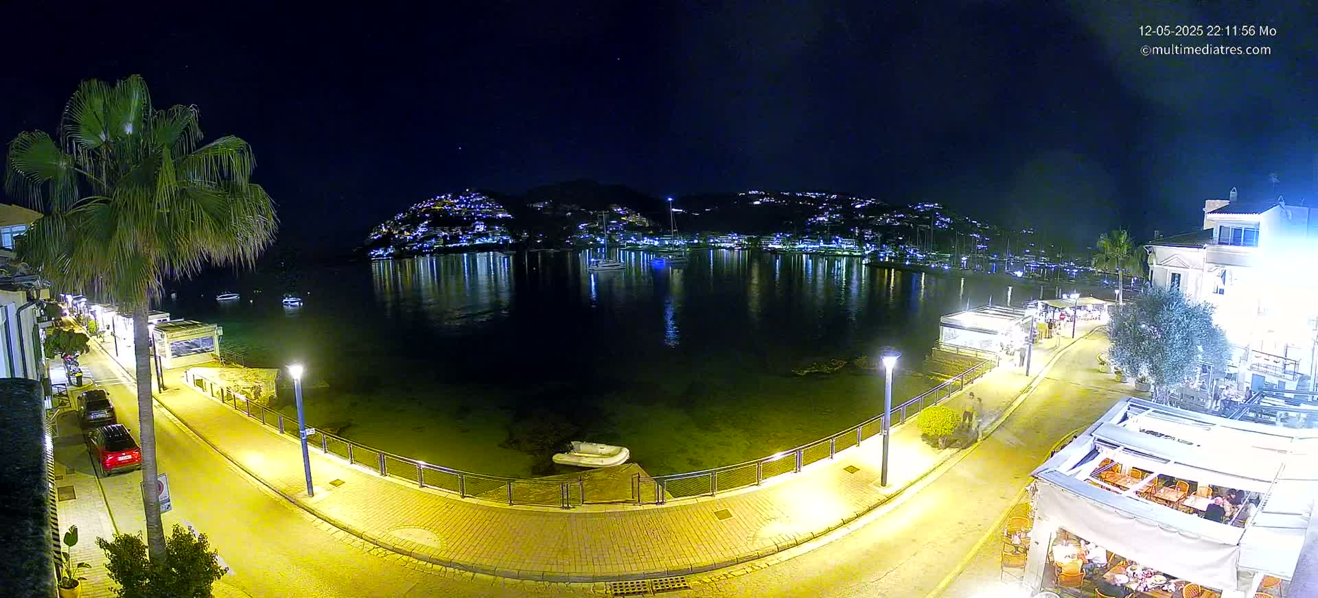 A nighttime, illuminated waterfront scene shows a calm bay with boats, reflecting lights from buildings on a hillside across the water, and a paved walkway with cars and outdoor seating at restaurants.