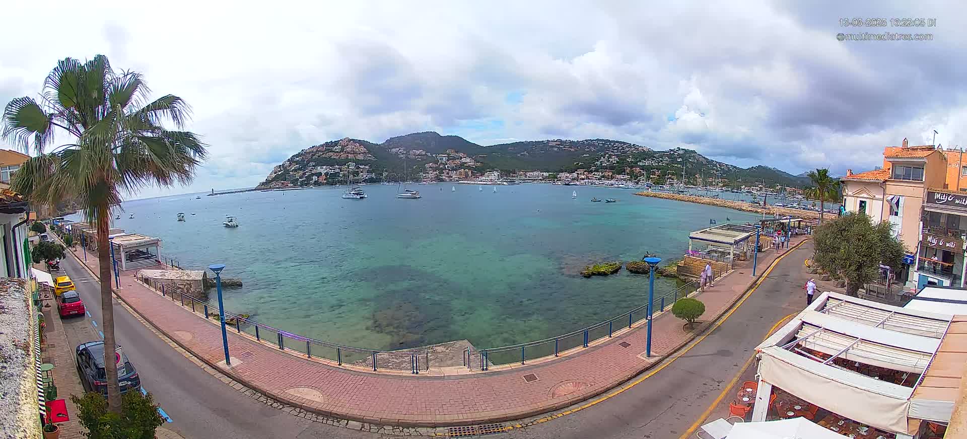 A partly cloudy day reveals a waterfront scene with a calm, clear, teal-colored bay filled with boats, a paved walkway, and buildings lining a hilly shoreline.
