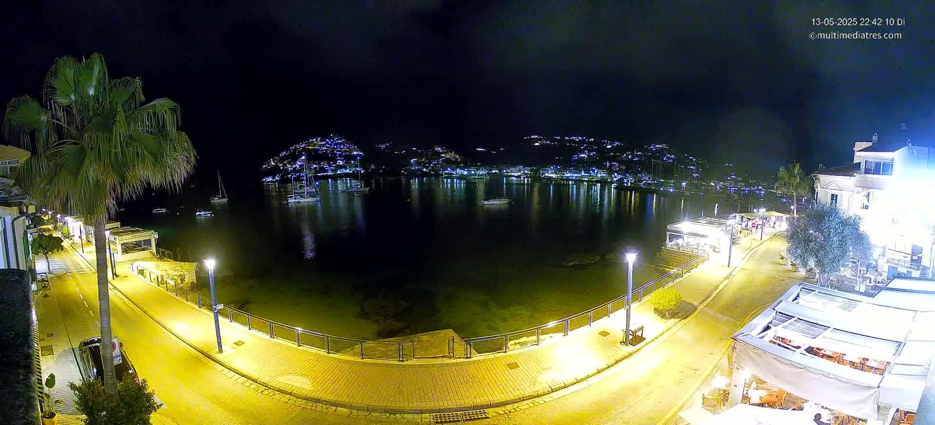 A nighttime view of a harbor town's waterfront, showing a calm body of water reflecting lights from buildings and boats, with a paved walkway and outdoor restaurants lining the shore.