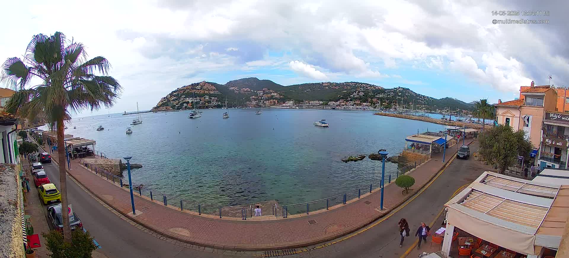 A seaside town with a calm bay filled with boats, under a partly cloudy sky, is seen from a high vantage point, showing a paved walkway and street leading to buildings and restaurants.