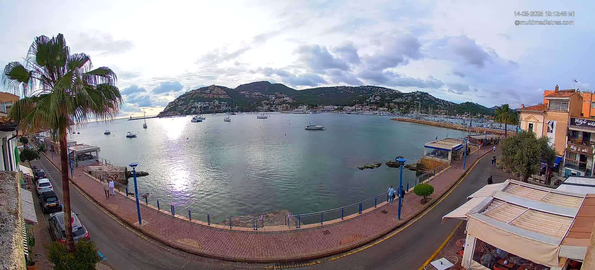A calm bay with numerous boats is viewed from a waterfront promenade under a partly cloudy sky.