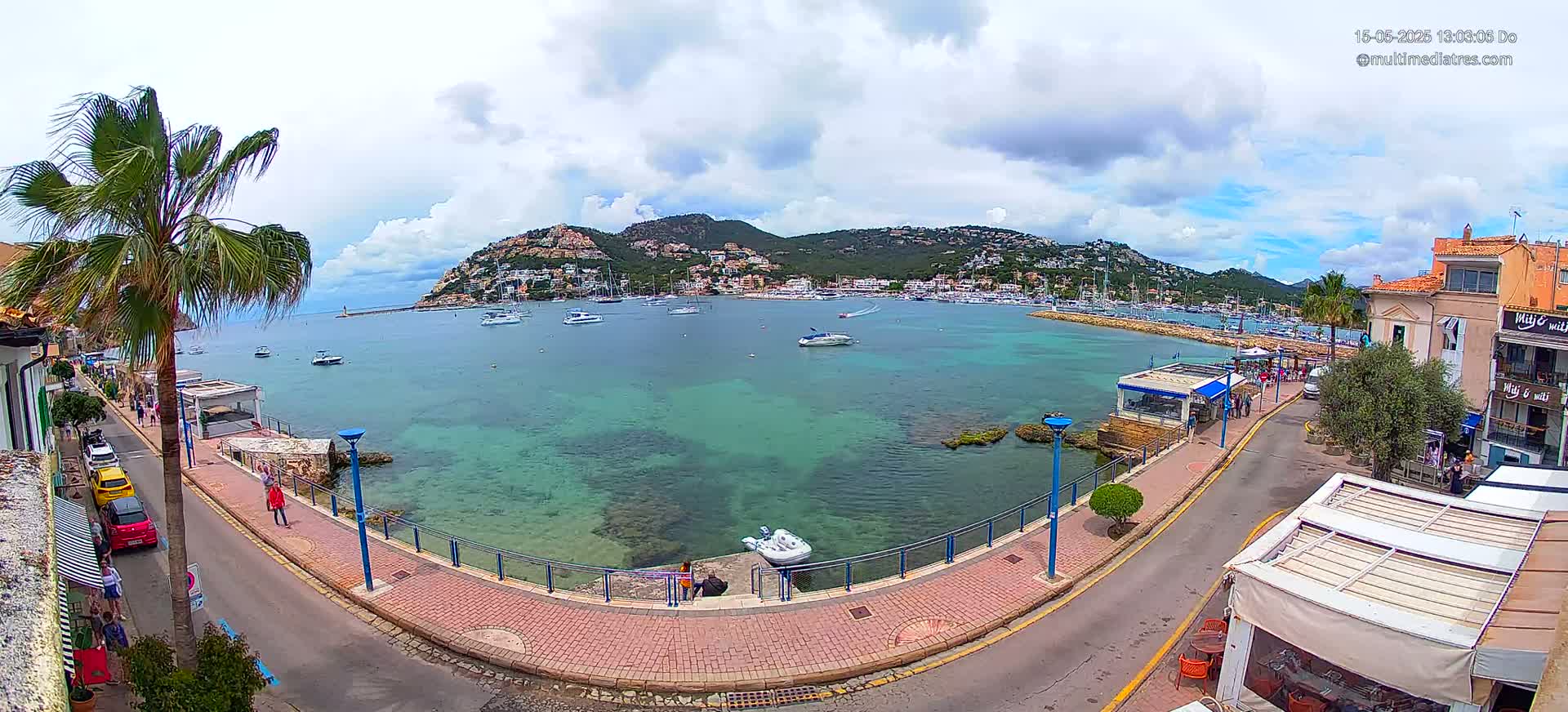 A partly cloudy day reveals a calm harbor filled with boats, nestled beside a paved waterfront walkway lined with buildings and palm trees, all set against the backdrop of a hillside town.
