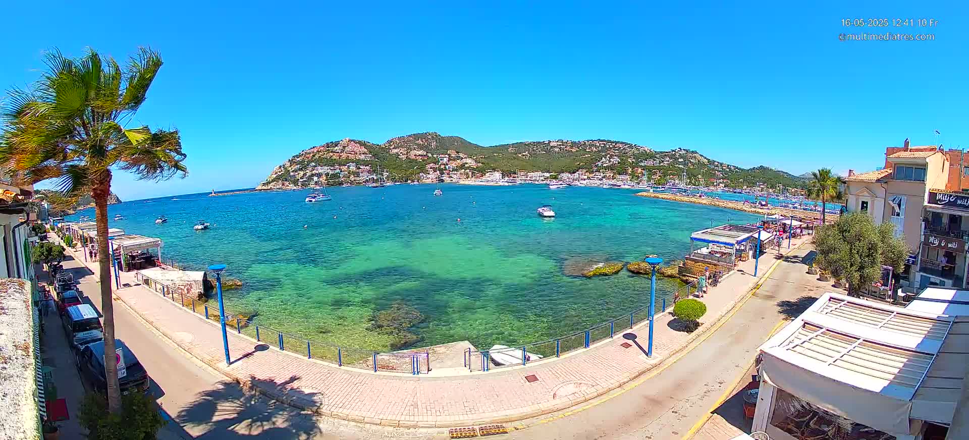A clear, sunny day shows a calm bay with clear, shallow turquoise water, boats, a seaside promenade with buildings and palm trees, and a hill covered with buildings in the background.