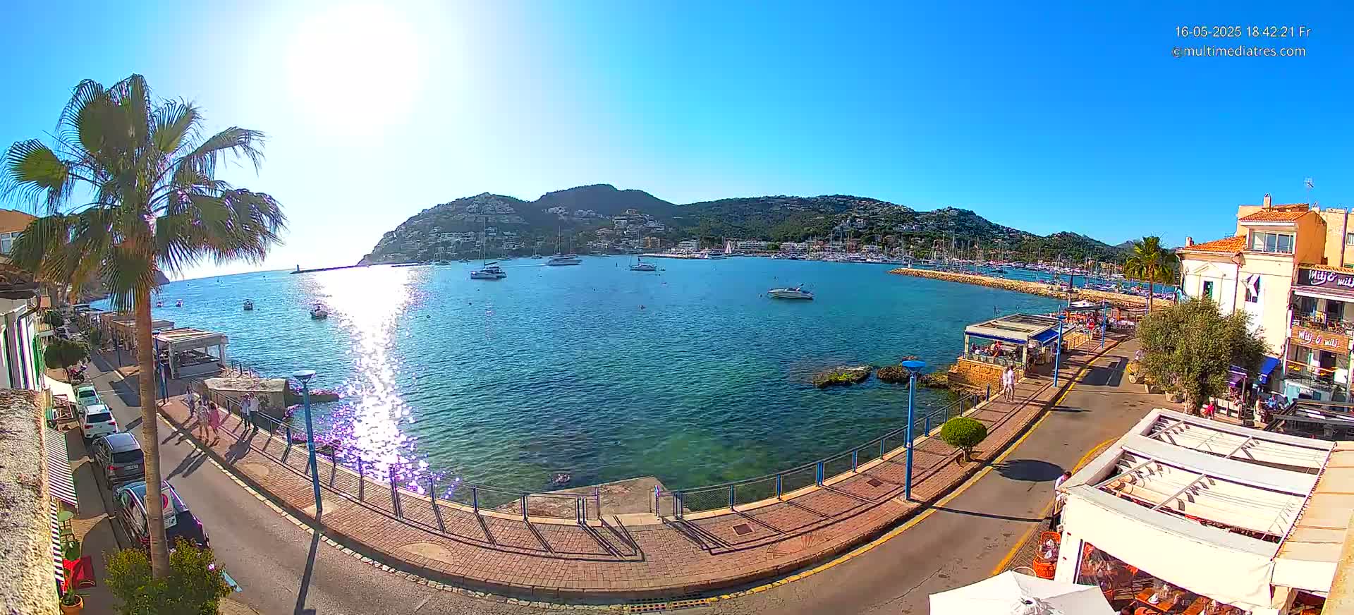A sunny seaside town with a calm harbor filled with boats, bordered by a paved walkway, restaurants, and buildings, nestled beneath a hill.