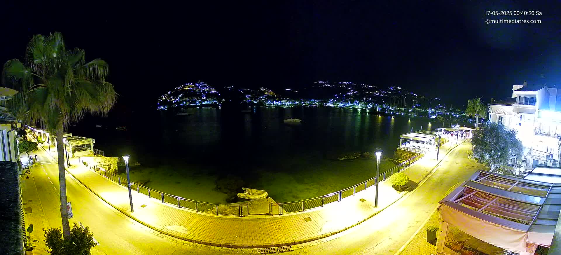 A nighttime view of a calm bay with a town's lights illuminating the hillsides across the water, a curving illuminated walkway along the shore, and a small boat near the water's edge.