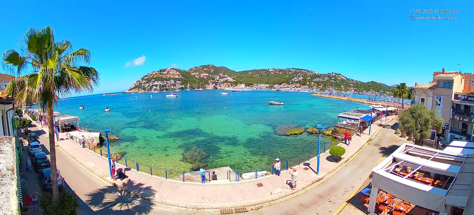 A clear, sunny day shows a coastal town with a calm, turquoise bay filled with boats, a paved walkway, and buildings lining the shore.