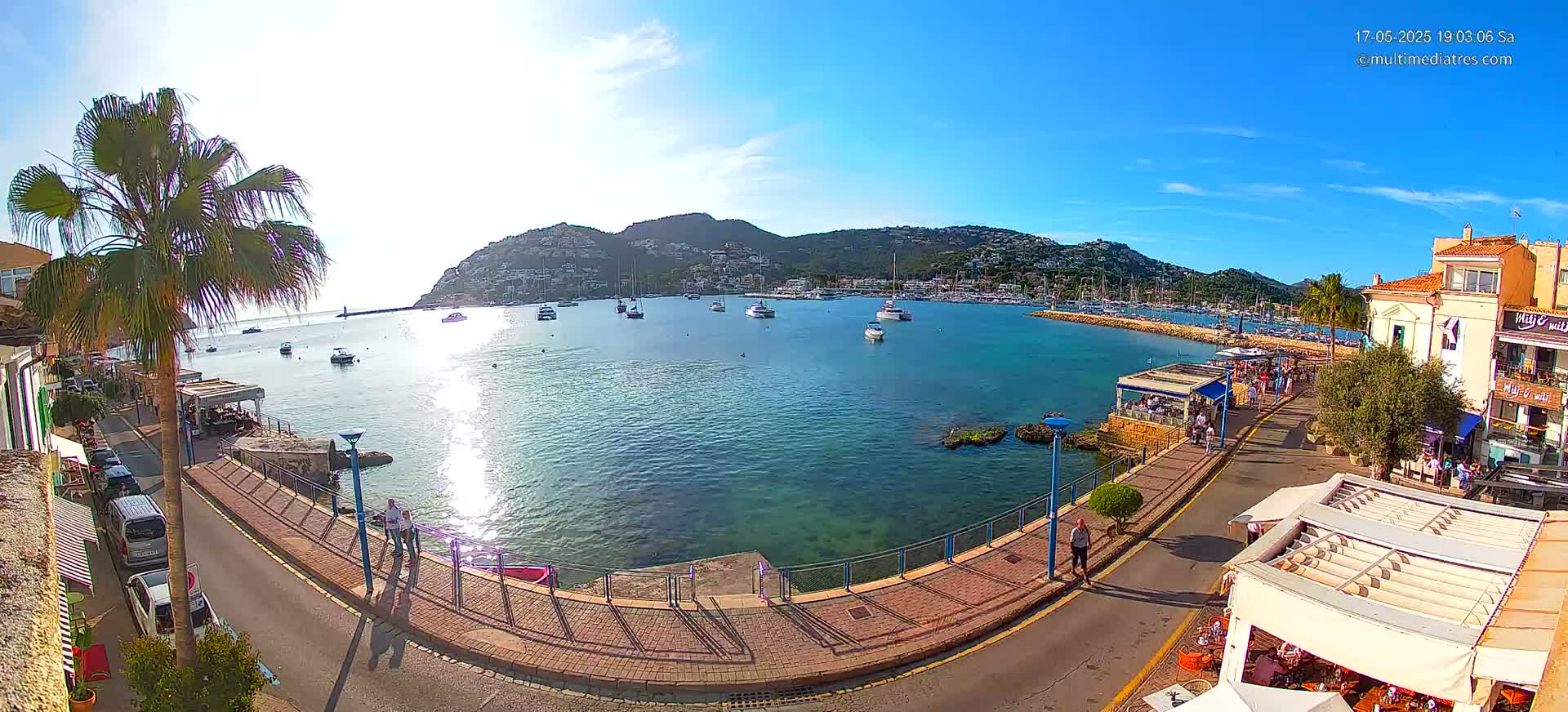 A sunny harbor scene shows a calm bay filled with boats, a waterfront promenade with restaurants and people, and a hill covered in buildings in the background.