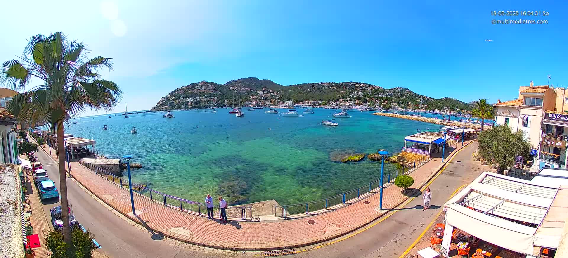 A sunny seaside town scene shows a calm, clear turquoise bay filled with boats, a paved walkway lined with buildings and restaurants, and a palm tree near the water.