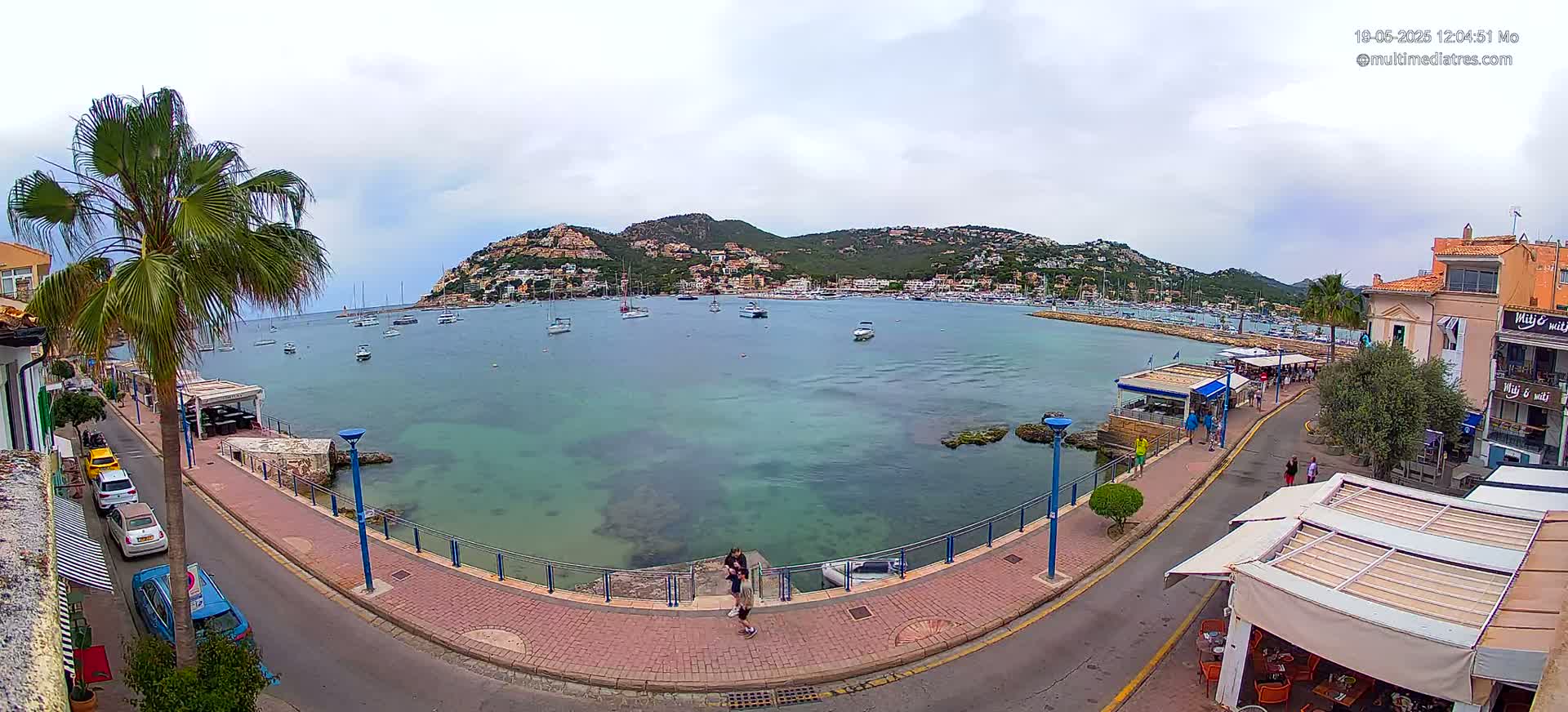 A calm bay with numerous boats is seen from a waterfront promenade on a partly cloudy day, with a hill covered in buildings in the background.