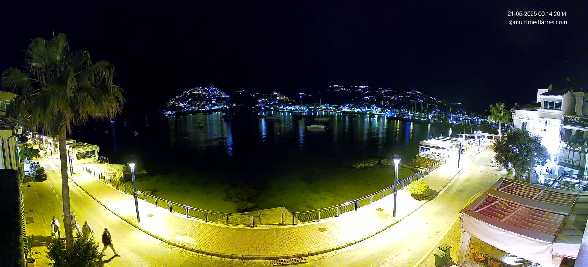 A nighttime view of a waterfront promenade, lit by streetlights, overlooking a calm bay with a hilltop town illuminated across the water.