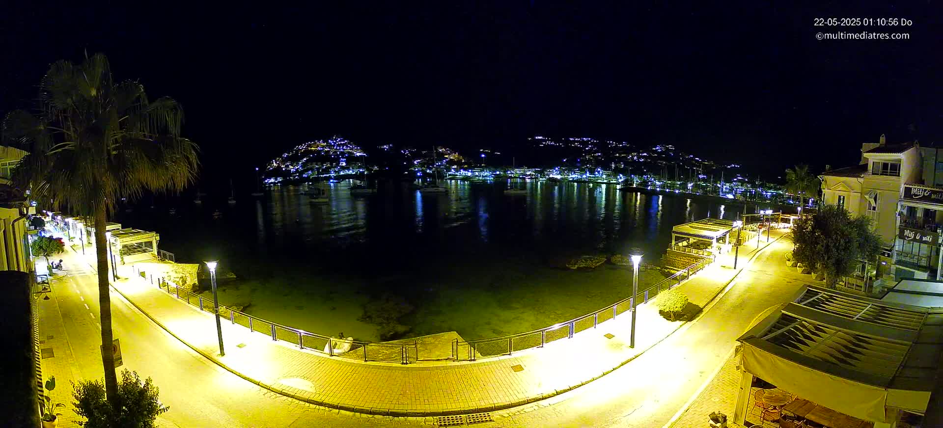 A nighttime view of a waterfront promenade illuminated by streetlights, overlooking a calm harbor filled with boats and a hill dotted with lights in the background.