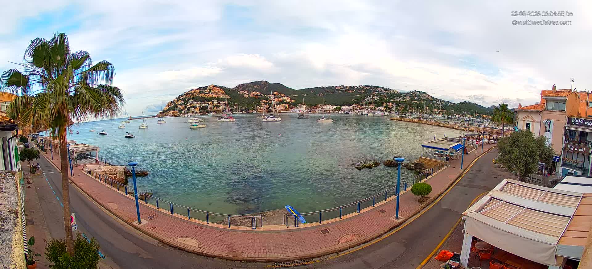 A mostly sunny day shows a calm harbor filled with sailboats, a waterfront promenade, and a hill covered in buildings in the background.