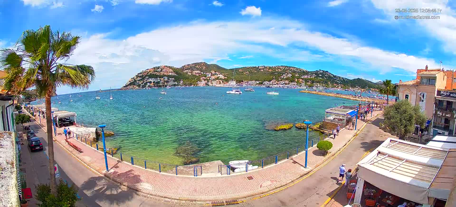 A sunny day overlooking a calm, clear-water harbor filled with boats, a waterfront promenade with buildings and people, and a hill covered in buildings in the background.