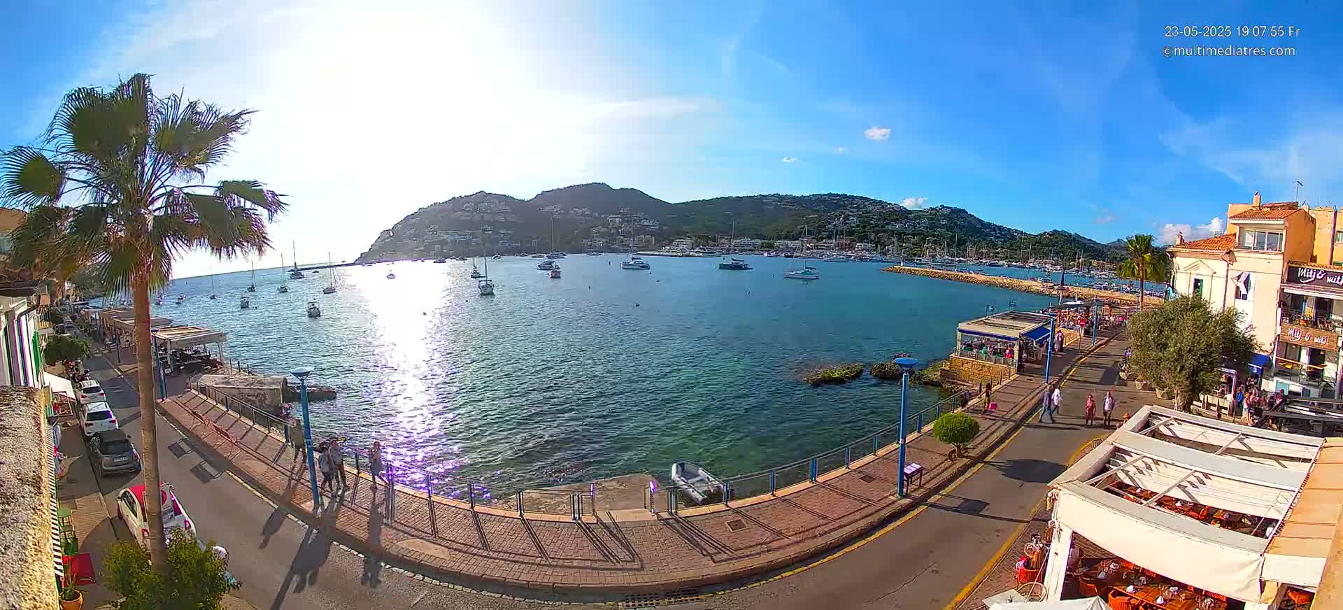 A sunny harbor scene shows a calm body of water filled with sailboats and yachts, nestled against a hillside town under a clear blue sky.
