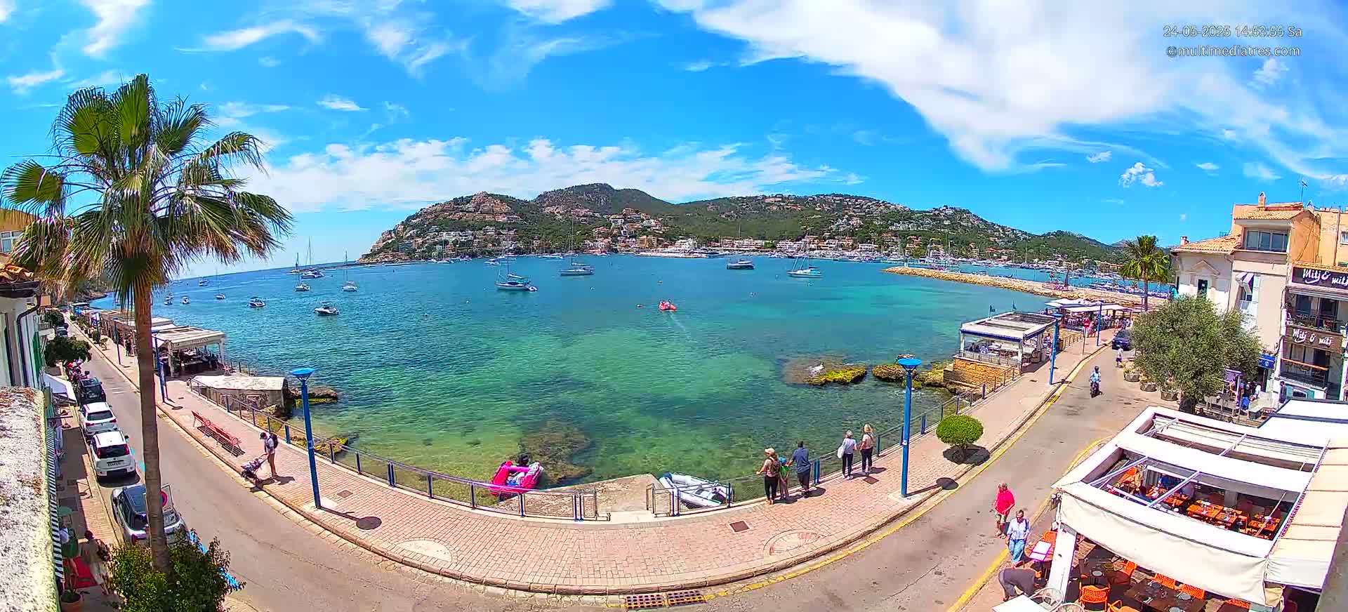 A sunny day in a harbor town shows clear blue water with boats, a paved walkway lined with buildings and people, and a hill in the background.