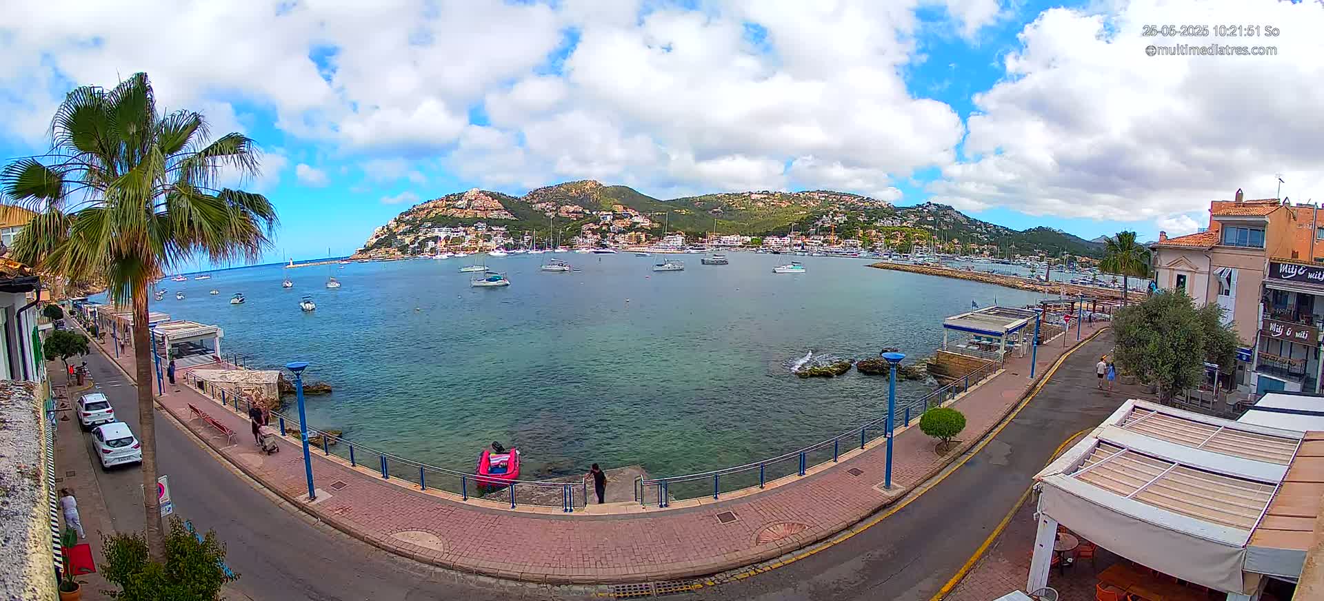 A partly cloudy day shows a seaside town with a calm bay filled with boats, a paved walkway, and palm trees, nestled against a hillside of buildings.