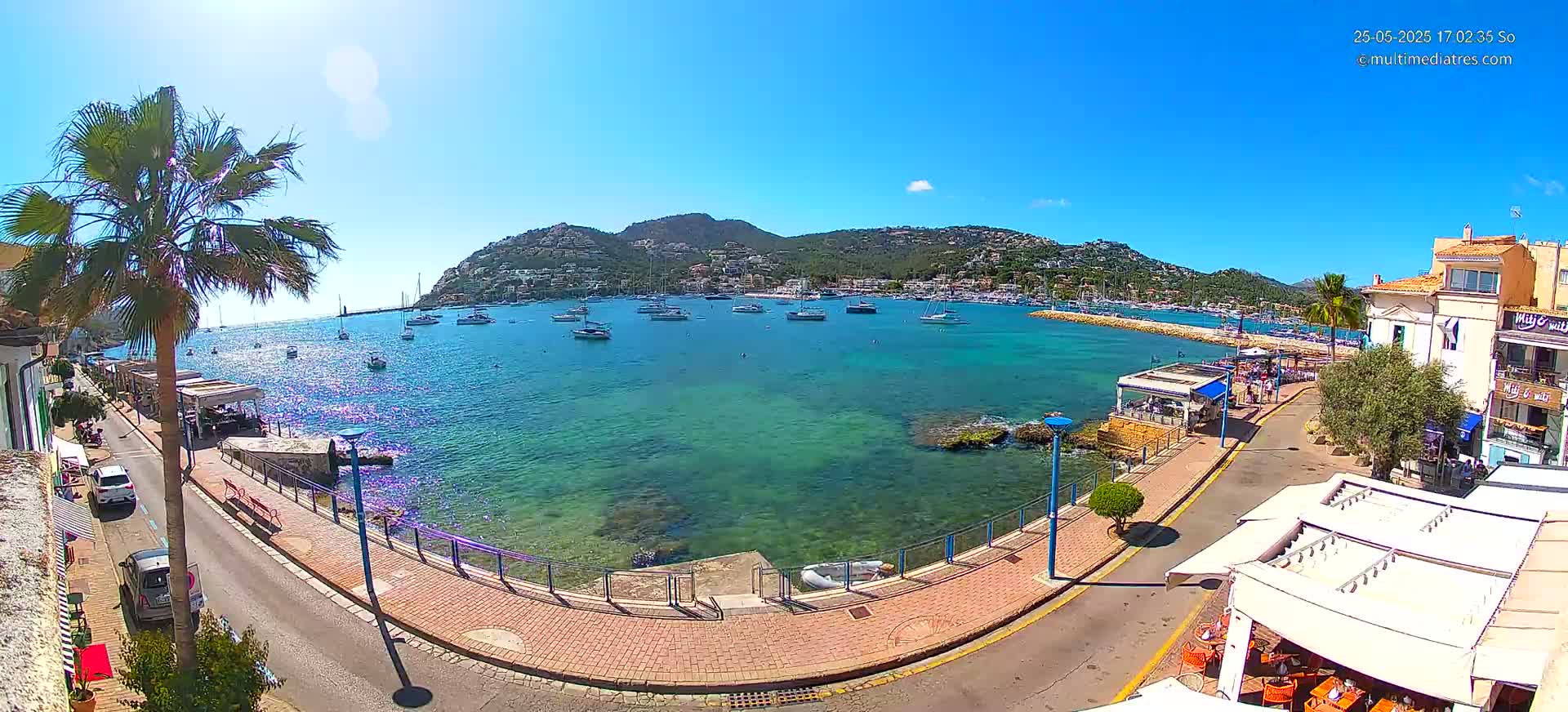 A sunny harbor scene shows a calm, clear, turquoise body of water filled with sailboats, a paved walkway lined with restaurants and palm trees, and a hillside town in the background.