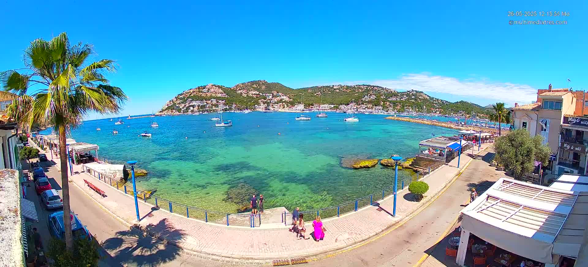 A sunny day reveals a clear, turquoise bay filled with boats, overlooked by a town nestled against a verdant hillside.