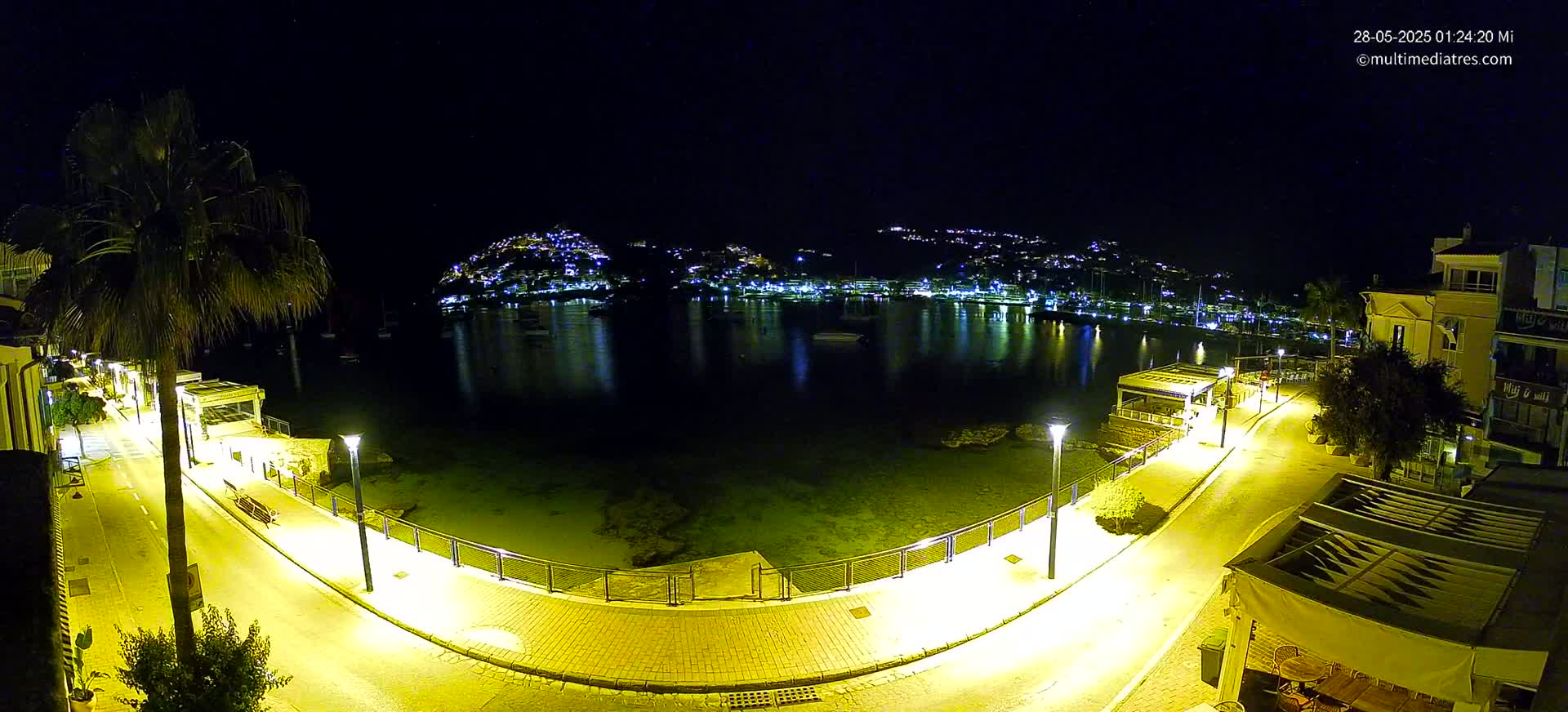 A nighttime, brightly lit seaside promenade curves around a calm bay, with a hilltop town twinkling with lights in the background under a clear, dark sky.