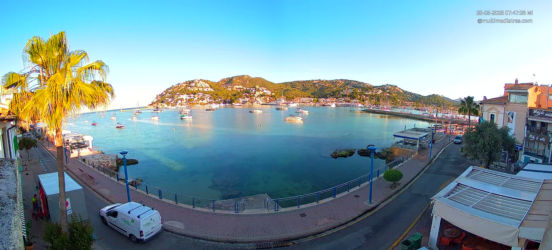 A clear, sunny day shows a waterfront scene with numerous boats in a calm bay, a paved walkway, and buildings along the shore, with palm trees visible.