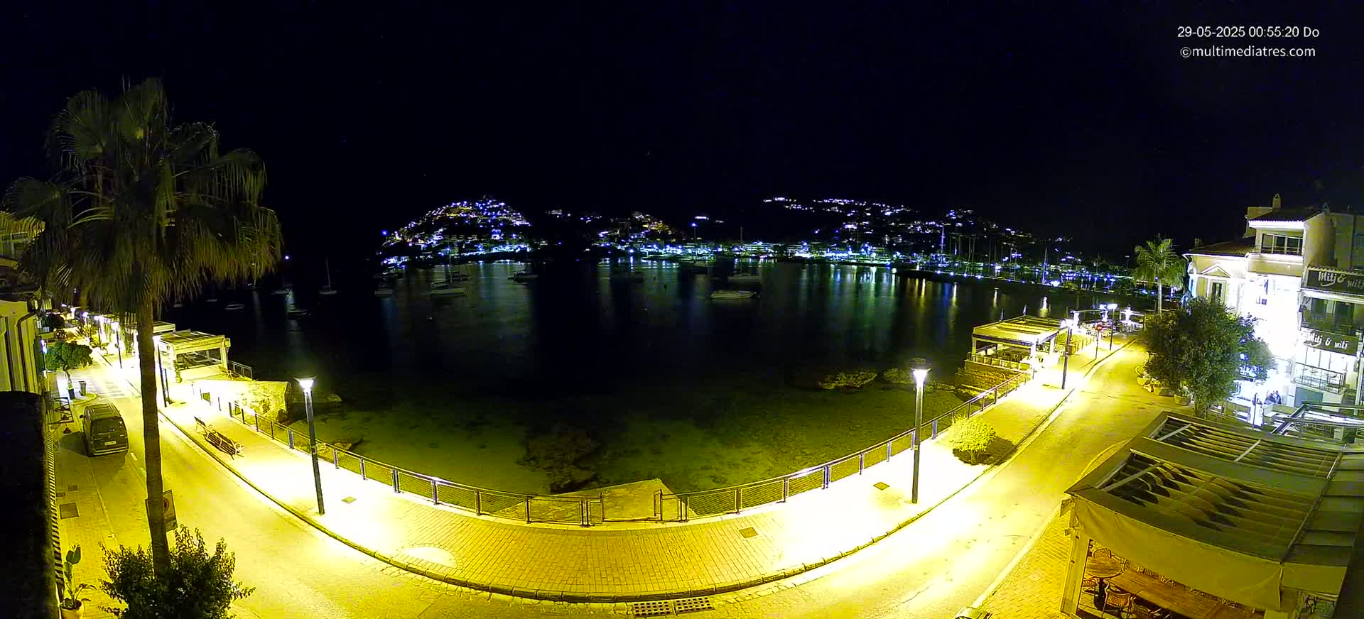A nighttime view of a calm harbor, illuminated by streetlights, with buildings and boats visible across the water.