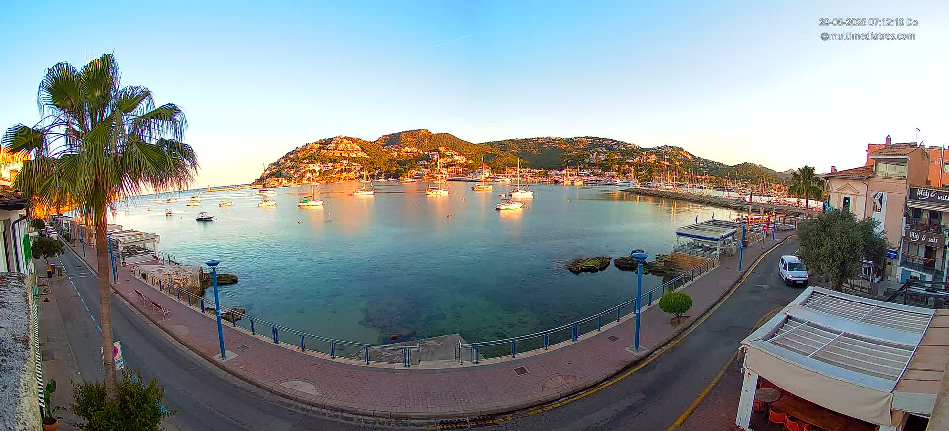 A calm bay filled with numerous boats is seen from a waterfront road on a clear, sunny morning, with a hill covered in buildings in the background.
