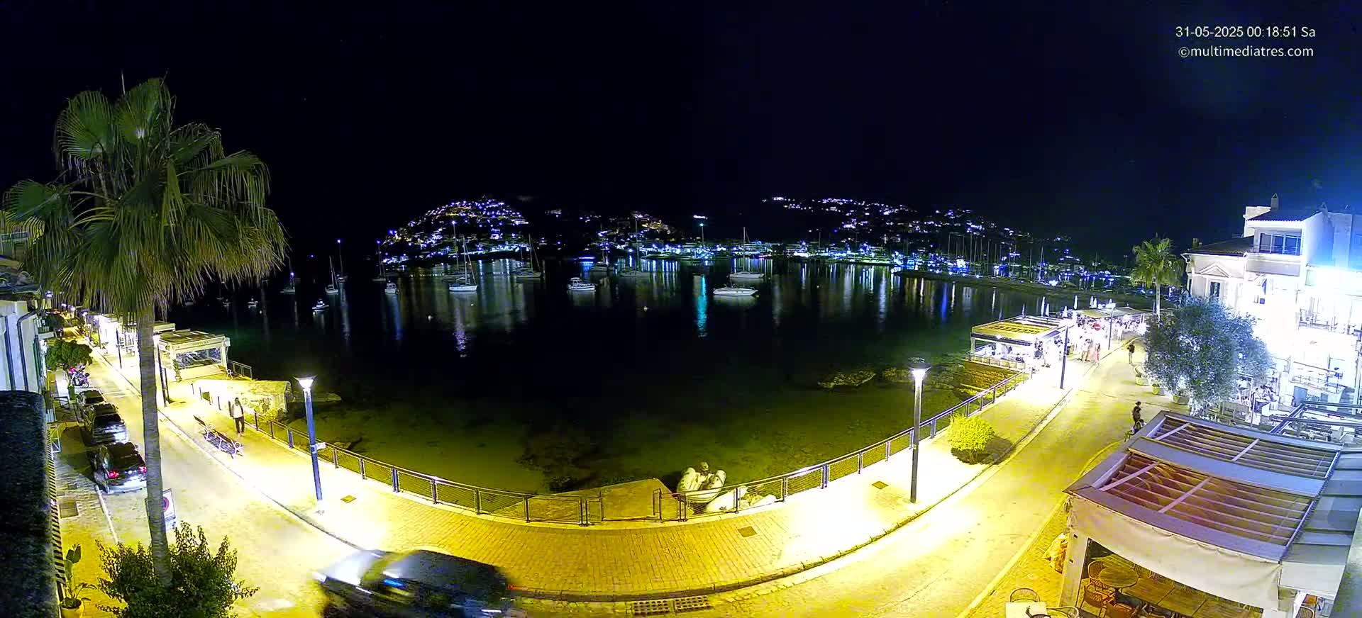 A nighttime view of a calm harbor filled with boats,  lined by a brightly lit waterfront promenade with people strolling, palm trees, and buildings.