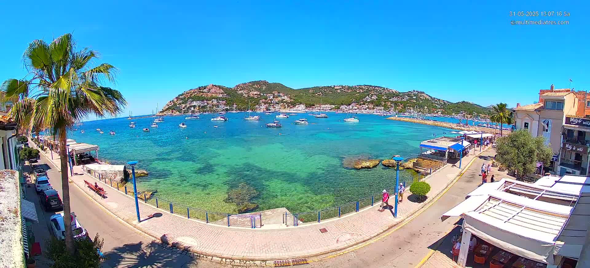 A clear, sunny day overlooking a calm, turquoise bay filled with boats, a town nestled against a hillside, and a paved walkway lined with buildings and people.