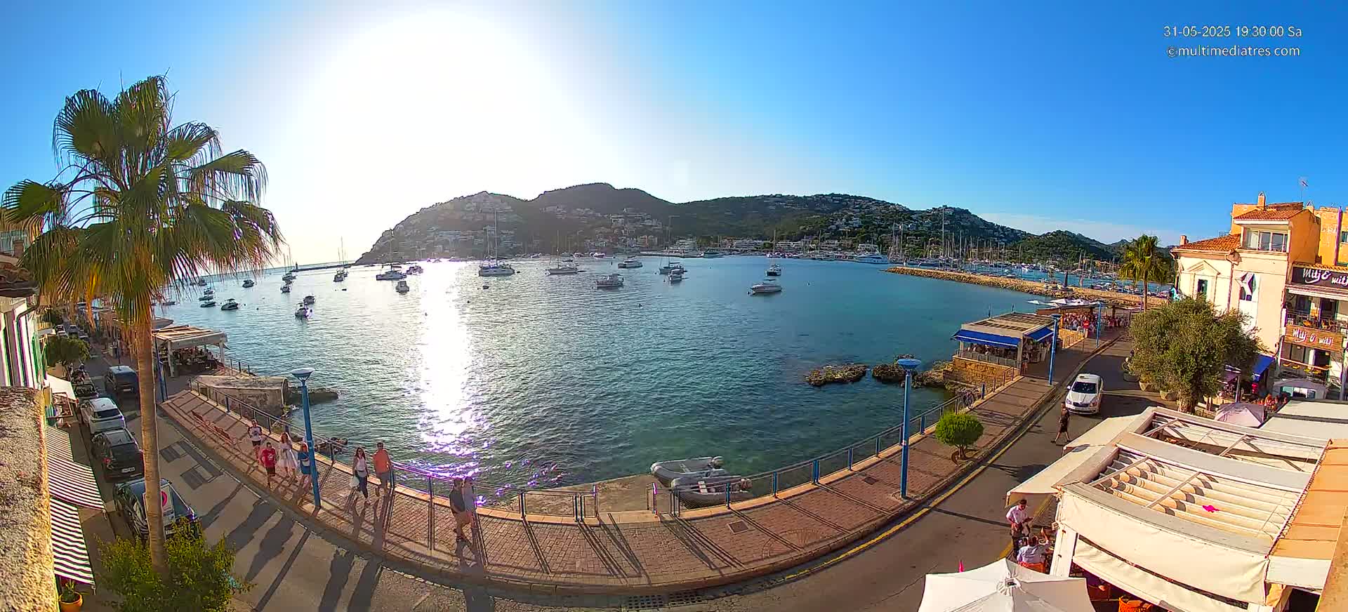 A sunny harbor scene shows numerous boats in calm water, a waterfront promenade with people walking, and buildings lining the shore against a backdrop of a hillside.