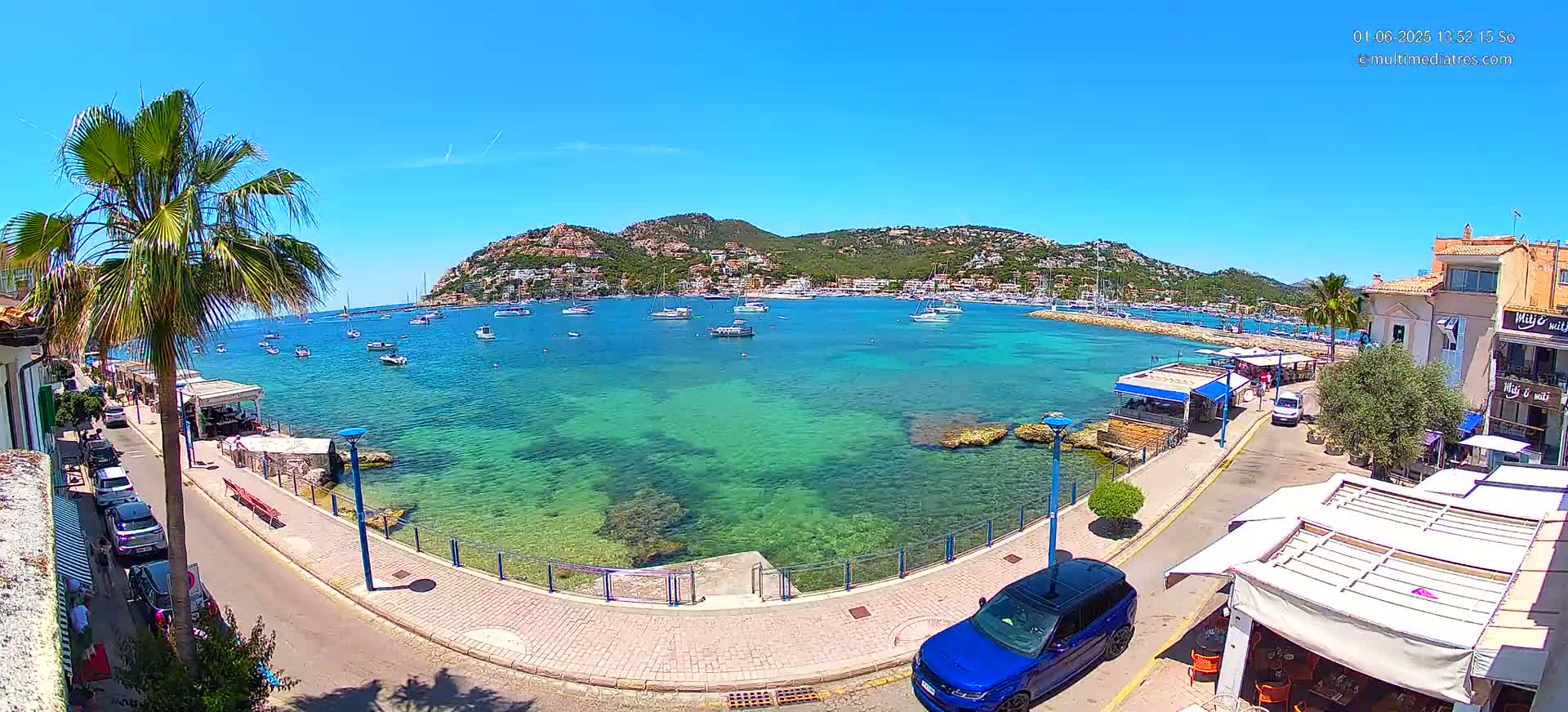 A sunny day overlooking a clear, turquoise harbor filled with boats, a waterfront promenade lined with buildings and palm trees, and a blue SUV parked nearby.