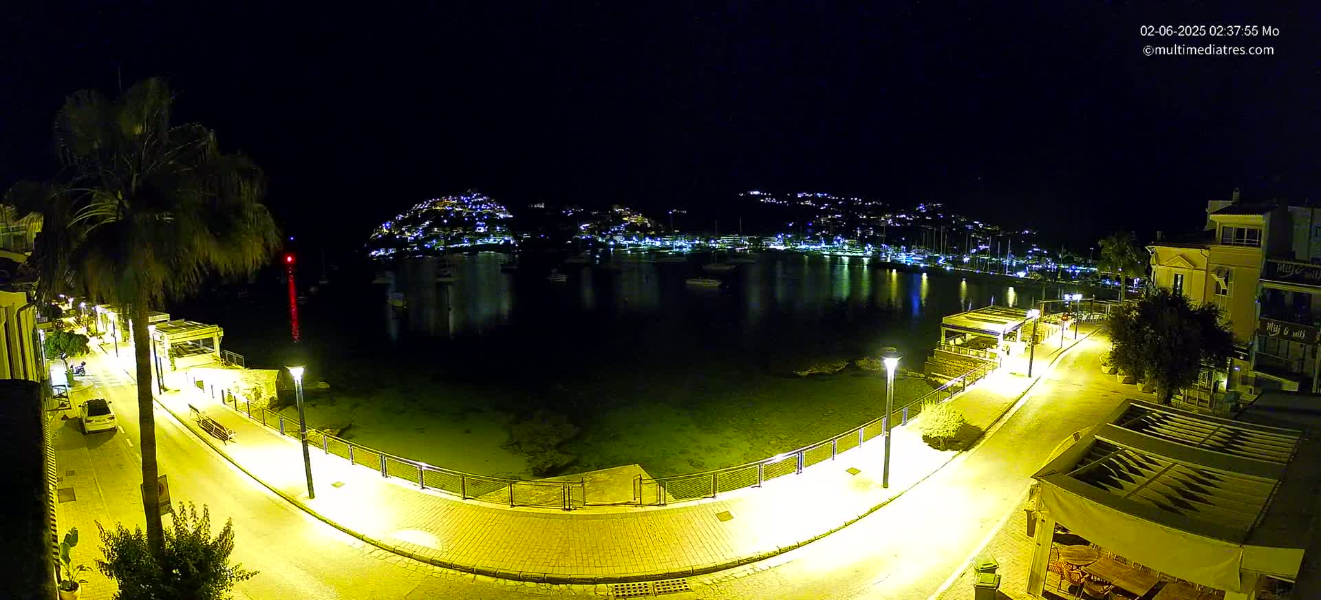 A nighttime, illuminated waterfront scene shows a calm bay with moored boats, a hilltop town twinkling with lights, and a curving paved walkway with lampposts lining a shoreline.