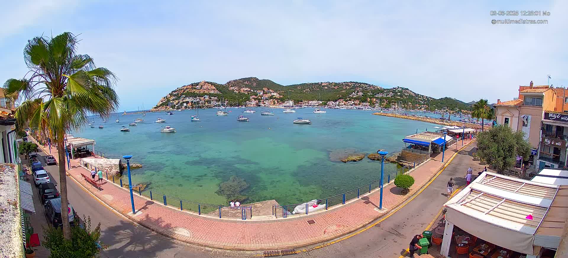 A seaside town on a sunny day with clear, shallow turquoise water, numerous boats in the harbor, and buildings lining a paved walkway.