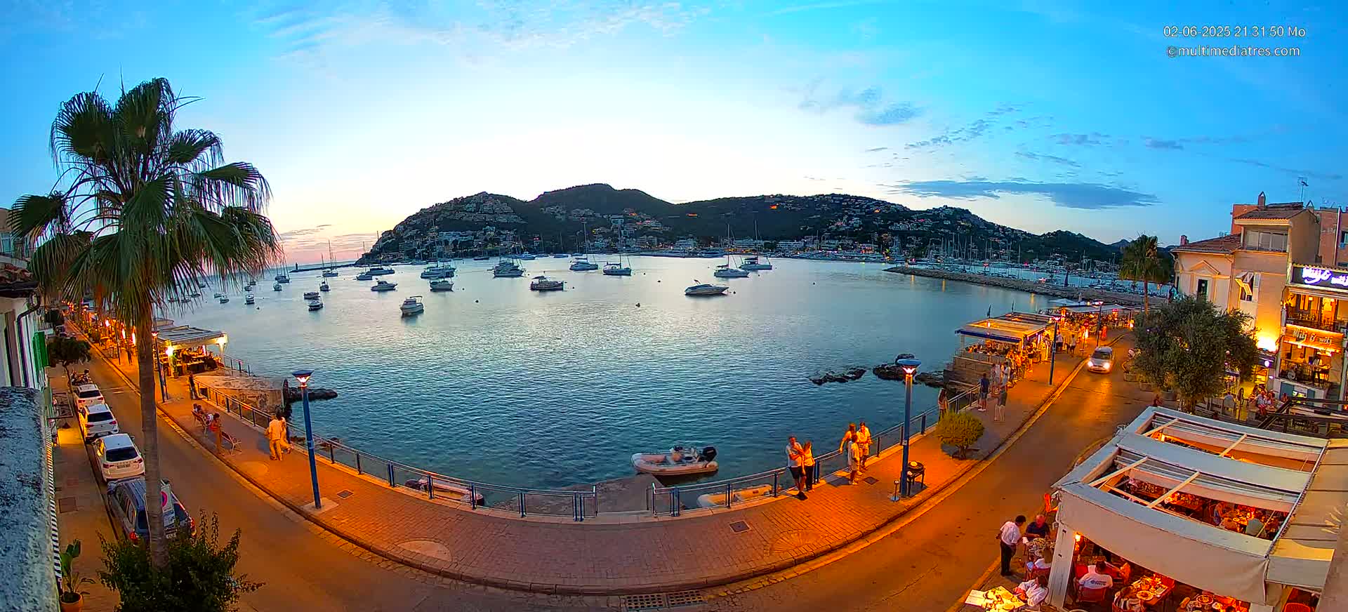 A waterfront town at dusk, with boats in the harbor and people strolling along a lit promenade under a clear sky.