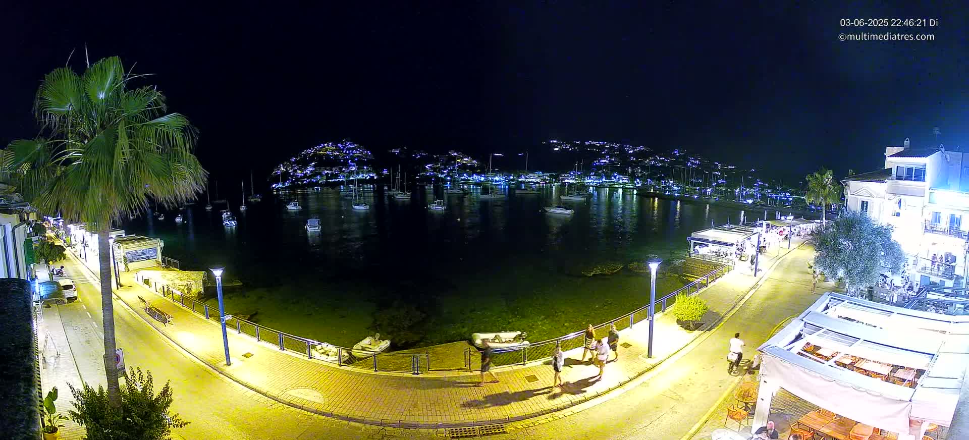 A nighttime, illuminated waterfront scene shows a calm harbor with moored boats, a paved walkway lined with people and buildings, and a hill in the distance with many lights.