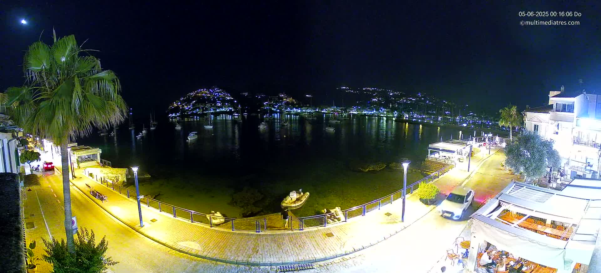 A nighttime aerial view of a calm harbor filled with boats, lined with illuminated buildings and a waterfront promenade under a clear, moonlit sky.