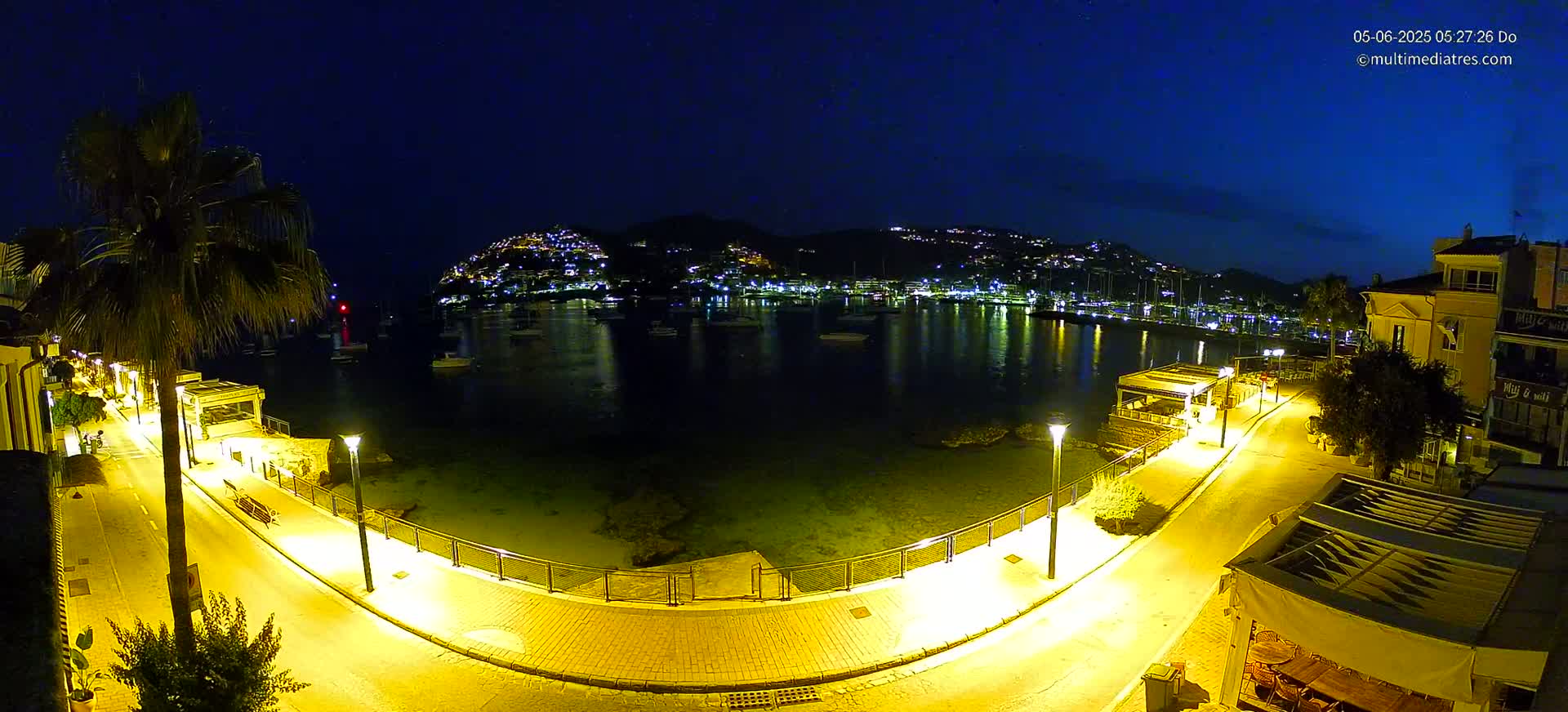 A nighttime view of a calm harbor illuminated by streetlights, with a hilltop town glowing in the distance under a dark blue sky.