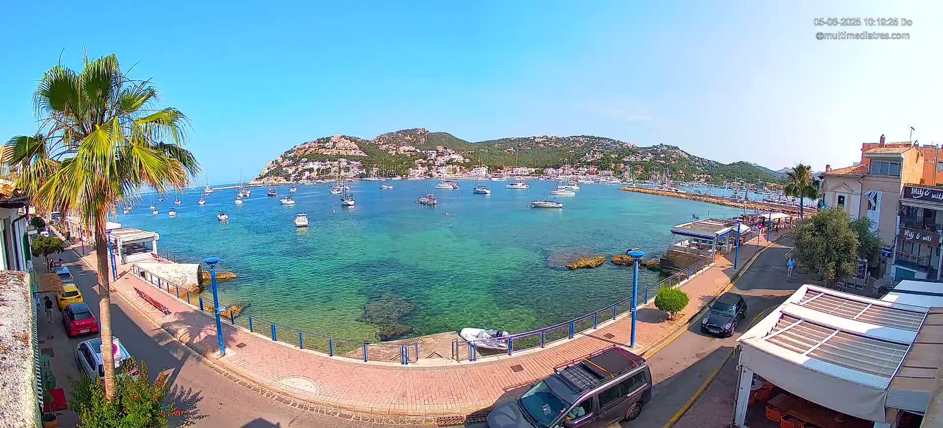 A clear, sunny day overlooks a calm, turquoise bay filled with sailboats and motorboats,  a waterfront promenade lined with buildings and palm trees.